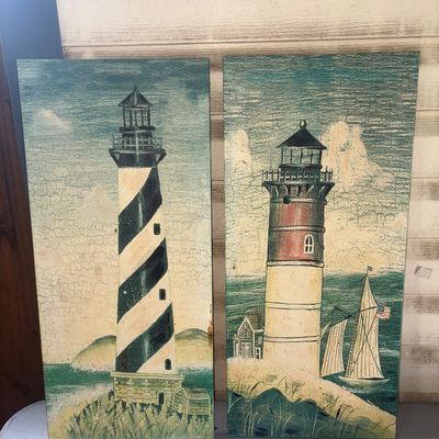 Sale Photo Thumbnail #372: Pair of decorative rectangular wooden wall art panels featuring painted lighthouse scenes with a distressed/crackle finish. One panel features a black and white spiral lighthouse, and the other shows a red and white lighthouse with a sailboat in the backg