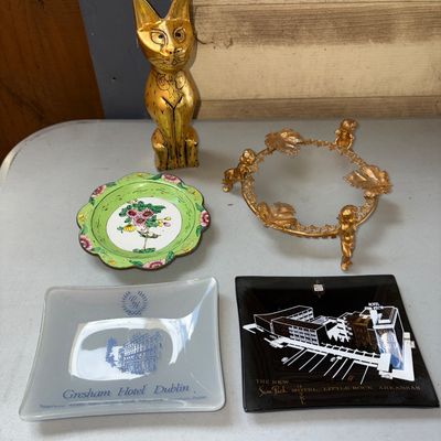 Sale Photo Thumbnail #40: An eclectic 5-piece collection including a styled gold-tone/brass carved standing cat figurine, a ornate gilt metal cherub/putti dish or globe stand with leaf detailing, a green floral enameled scalloped dish, a glass Gresham Hotel Dublin souvenir dish, a