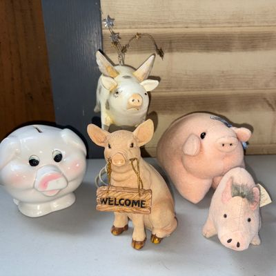Sale Photo Thumbnail #32: A collection of 5 pig-themed decorative items including a ceramic piggy bank with coin slot, a resin seated pig figurine holding a "WELCOME" sign, a winged/horned spotted pig figure with star wire accents, and two small plush soft pig figures. Items show 