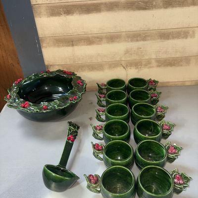 Sale Photo Thumbnail #337: A ceramic holiday punch bowl set featuring a green glazed finish with raised holly leaf and red berry accents around the rims and handles. Includes 1 large punch bowl, 1 ceramic ladle, and 12 matching cups. Minor wear visible, including a small chip on on