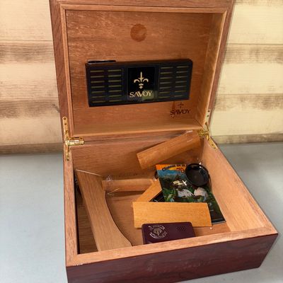 Sale Photo Thumbnail #104: Savoy desktop cigar humidor featuring a rich reddish-brown polished wood finish and Spanish cedar lining. Inside includes an attached Savoy humidifier element, loose wooden divider pieces, and various small accessories. Shows minor surface dust and light 