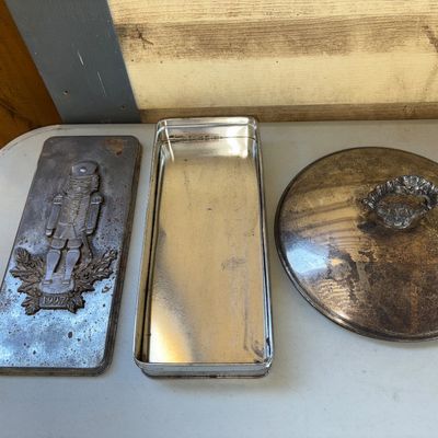 Sale Photo Thumbnail #223: Lot includes a vintage rectangular metal tin dated 1997 with an embossed nutcracker design on the lid, along with a round silver-tone metal lid featuring an ornate decorative handle. Both items show signs of age including surface oxidation, tarnishing, ru