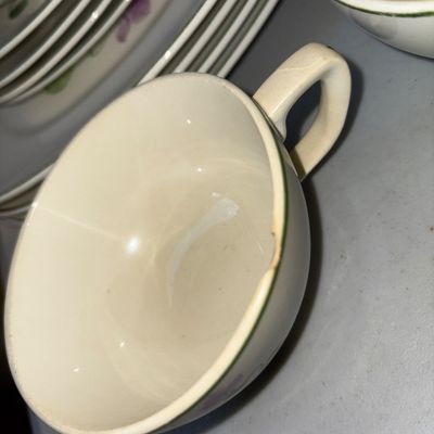 Sale Photo Thumbnail #305: A set of vintage cream-colored ceramic dinnerware featuring a hand-painted or printed purple floral violet pattern with green trim. The lot includes dinner plates, small bowls/saucers, a large serving platter, and 4 teacups. Items show typical vintage wea