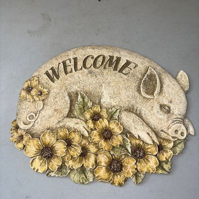 Sale Photo Thumbnail #364: A decorative outdoor or garden welcome plaque featuring a sleeping pig surrounded by yellow flowers. Made of molded resin or cast stone material with textured and painted details. Appears to be in good used condition with light surface wear.
10.5X8.5in