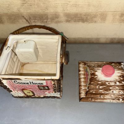 Sale Photo Thumbnail #181: Vintage ceramic cottage-style cookie jar labeled 'Cookie House'. Features a painted log cabin design with pink accents, a removable roof lid, and a woven wicker overhead handle. Shows some visible crazing, minor wear, and a hairline crack visible along th