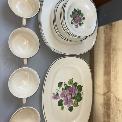 Sale Photo Thumbnail #304: A set of vintage cream-colored ceramic dinnerware featuring a hand-painted or printed purple floral violet pattern with green trim. The lot includes dinner plates, small bowls/saucers, a large serving platter, and 4 teacups. Items show typical vintage wea