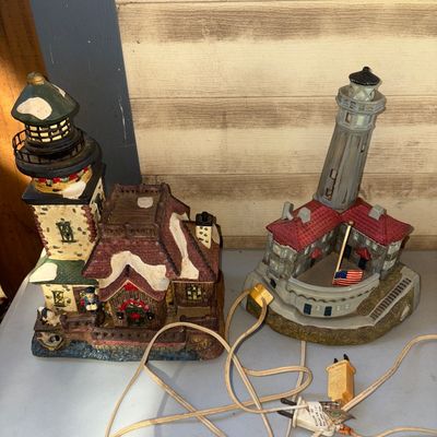 Sale Photo Thumbnail #275: Set of two miniature lighthouse building figurines with power cords, including a snowy winter holiday style lighthouse and a coastal lighthouse with an American flag. Both feature detailed painted resin construction. Light bulbs and cords are attached; li