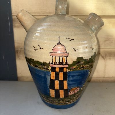 Sale Photo Thumbnail #350: A glazed ceramic/pottery botijo or dual-spout water jug featuring a hand-painted coastal scene with a checkered lighthouse, water, shore, and birds. Includes a top handle, pouring spout, and filling spout. Appears to be in good pre-owned condition with no
