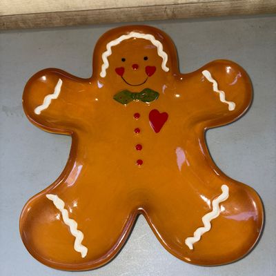 Sale Photo Thumbnail #216: Glazed ceramic gingerbread man serving platter or decorative dish, featuring hand-painted details including a green bow tie, red heart, and white icing trim. Appears to be in good condition with no visible chips or cracks.
10X9.5in