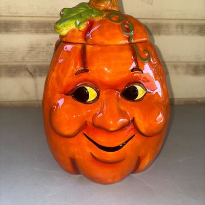 Sale Photo Thumbnail #155: Ceramic or pottery pumpkin cookie jar featuring a smiling jack-o'-lantern face and a removable lid styled with a stem and green leaf detail. Bright orange glazed finish with a white interior. Appears in good pre-owned condition with no visible chips or ma
