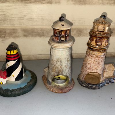 Sale Photo Thumbnail #144: A group of three lighthouse-themed decorative items, including one painted resin or ceramic lighthouse figurine featuring black and white stripes, and two ceramic/stoneware tea light candle holders styled as detailed lighthouses. Items show light surface 