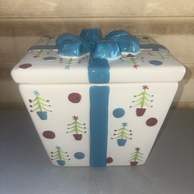 Sale Photo Thumbnail #237: Ceramic cookie jar or covered vessel shaped like a wrapped present, featuring a hand-painted style Christmas tree and polka dot motif on a white background, topped with a dimensional blue ceramic bow lid. Appears to be in good condition with no visible ch