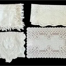 Collection of vintage white linens and lace items, including a fringed shawl, embroidered placemat, crocheted doily, and scalloped runner.