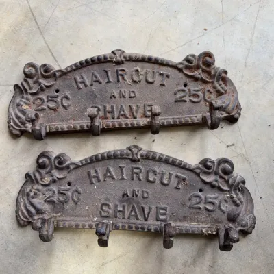 Sale Photo Thumbnail #245: Pair of vintage-style cast iron coat racks featuring embossed text that reads "HAIRCUT AND SHAVE 25¢". Each wall-mounted rack includes four hooks and exhibits a rustic, weathered patina.