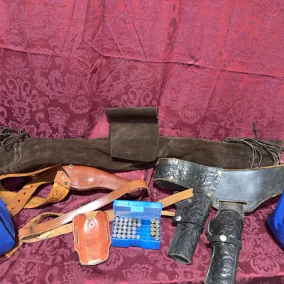 Sale Photo Thumbnail #60: Collection of Western shooting gear including tool-embossed leather gun belts with holsters, a brown suede rifle scabbard with fringe, blue nylon cases, and a box of ammunition.