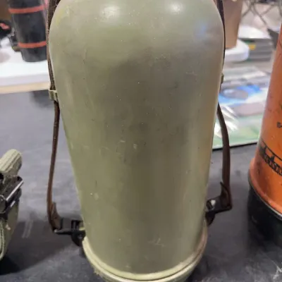 Sale Photo Thumbnail #51: Vintage military olive drab metal thermos with a brown leather carrying strap. Shows mild surface wear and scuffs consistent with age.
