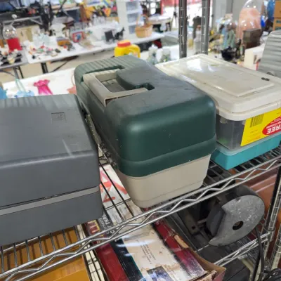 Sale Photo Thumbnail #217: Vintage Plano green and beige hard plastic fishing tackle box sitting on a metal shelving unit among other storage containers.