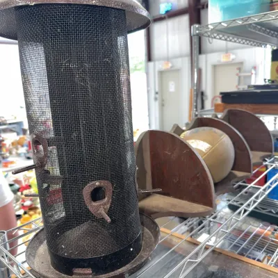 Sale Photo Thumbnail #212: Tall mesh metal bird feeder with feeding perches, sitting next to a wooden shelf unit on a wire rack.