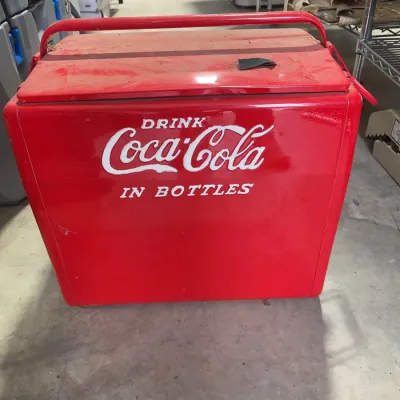 Sale Photo Thumbnail #170: Vintage red metal Coca-Cola cooler featuring embossed white lettering that reads "Drink Coca-Cola in Bottles" with a carrying handle.