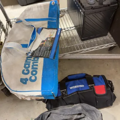 Sale Photo Thumbnail #233: Collection of storage and utility items including a vintage Sportcraft 4 Game Combo set bag, a black 12V car battery, a blue Workpro tool bag, and a dusty canvas duffel bag.