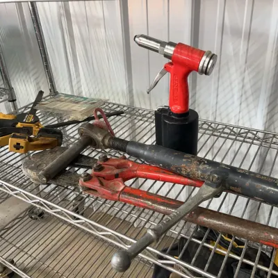 Sale Photo Thumbnail #28: Assorted heavy-duty hand and pneumatic tools including a red pneumatic rivet gun, bolt cutters, a ratcheting load binder, and bar clamps arranged on a metal wire shelf.
