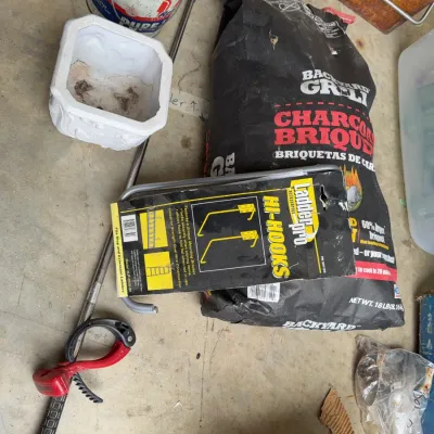 Sale Photo Thumbnail #252: Assorted household and garage supplies, including a bag of Backyard Grill charcoal briquettes, Ladder Pro hi-hooks in original packaging, a white planter, and a golf club.