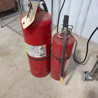 Sale Photo Thumbnail #13: Pair of vintage red fire extinguishers showing surface wear, dust, and inspection tags.