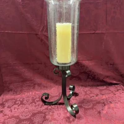 Sale Photo Thumbnail #14: Tall wrought iron hurricane candle holder featuring scrollwork legs and a clear glass cylinder, shown with a cream pillar candle.