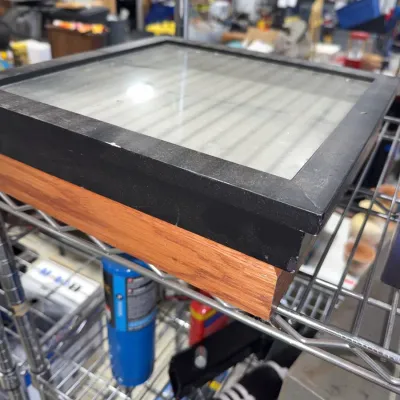 Sale Photo Thumbnail #215: Square wooden shadow box frame featuring a black outer bezel and glass top, resting on a wire rack.