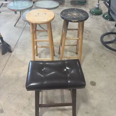 Sale Photo Thumbnail #67: A modern black faux-leather tufted vanity bench paired with two vintage wooden barstools showing surface wear and heavy paint distressing.