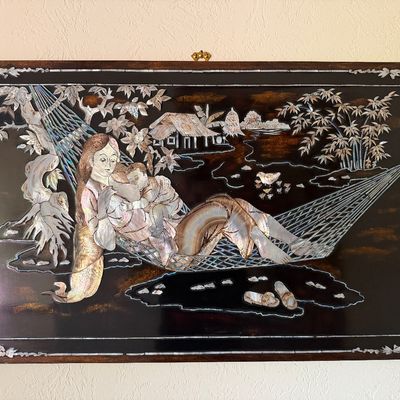 Vietnamese mother-of-pearl inlay lacquer wall plaque depicting a mother nursing a child in a hammock, set within an ornate decorative...