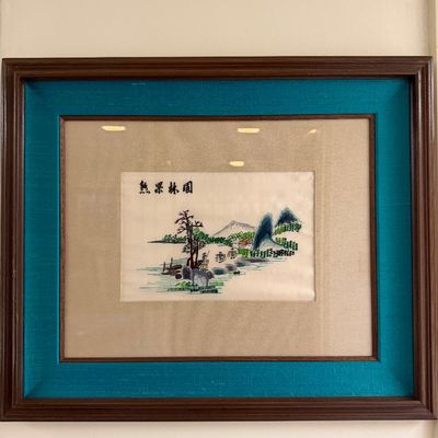 Framed Chinese silk embroidery artwork depicting a landscape with figures, mounted under glass with a teal inner mat and dark wood frame.