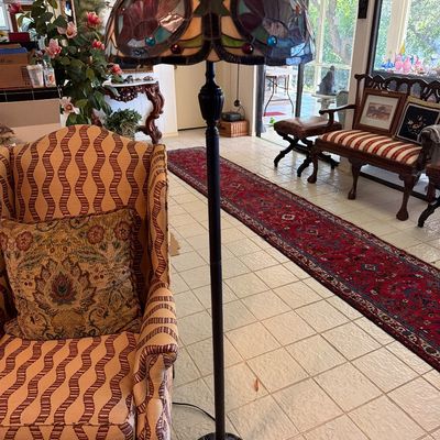 Tiffany-style leaded stained glass floor lamp with a dark metal pole and fluted base, set next to a patterned wingback armchair.