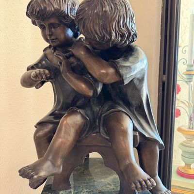 Bronze sculpture depicting two children seated on a bench, displayed on a green marble pedestal base.