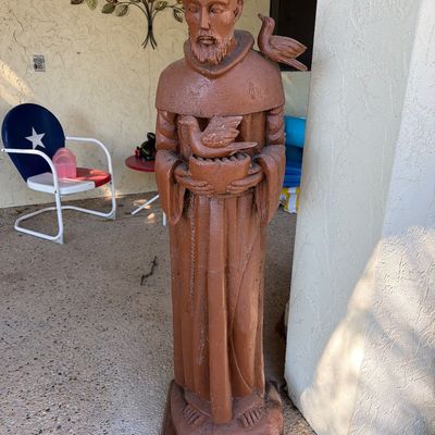 Large carved wooden outdoor statue of Saint Francis of Assisi depicting him holding a bird in a nest with a bird perched on his shoulder.