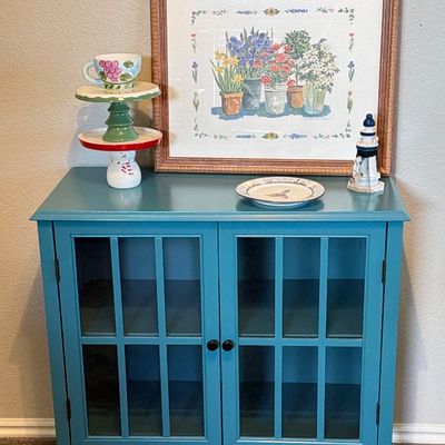 Teal accent cabinet featuring glass-paneled double doors, styled with framed floral wall art and decorative tabletop accents.
