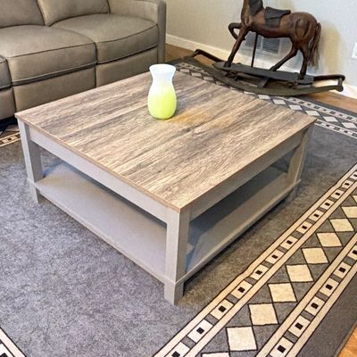 Square modern farmhouse-style coffee table with a weathered wood-grain top and lower shelf, accompanied by a geometric area rug, recliner...