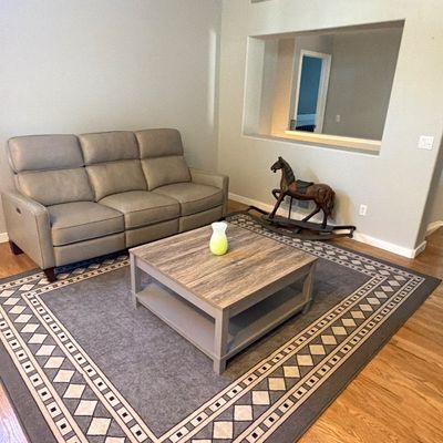 Living room setup featuring a gray leather reclining sofa, square wood-look coffee table, large geometric border area rug, and a vintage...