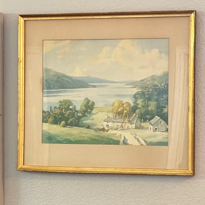 Framed watercolor landscape print depicting cottages along a scenic lake, presented in a mat and gold gilt wooden frame.