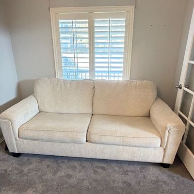 Contemporary cream-colored upholstered sofa featuring deep seat cushions and rolled arms, set against a window with plantation shutters.