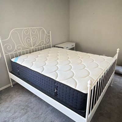 White metal scrollwork queen bed frame fitted with a thick quilted mattress and foundation, presented alongside a matching white...