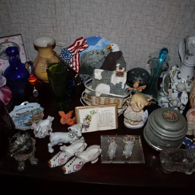 Sale Photo Thumbnail #150: A collection of decorative collectibles and glassware, including ceramic figurines, cobalt and cranberry glass vases, porcelain shoes, small trinket boxes, and a Madonna and Child statue.