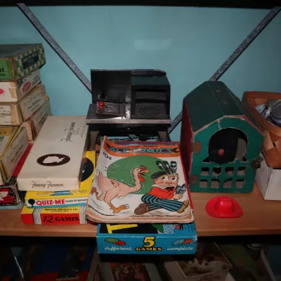 Sale Photo Thumbnail #70: Collection of vintage board games, puzzles, an Ace Comics issue, a miniature metal toy stove, and a wooden doghouse structure on a wooden shelf.
