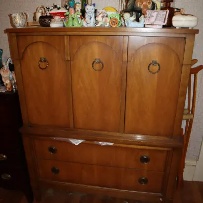 Sale Photo Thumbnail #147: Vintage mid-century wooden armoire dresser featuring arched panel doors with ring pulls over two wide drawers, topped with an assortment of decorative figurines and glassware.