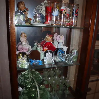 Sale Photo Thumbnail #115: Wooden curio cabinet filled with collectible figurines, decorated drinking glasses, small mirror displays, and green glass drinkware.