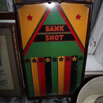 Sale Photo Thumbnail #78: Vintage 'Bank Shot' wooden Bagatelle pinball game board featuring vibrant green, yellow, and red graphics, displayed alongside framed artwork and toy train tracks.