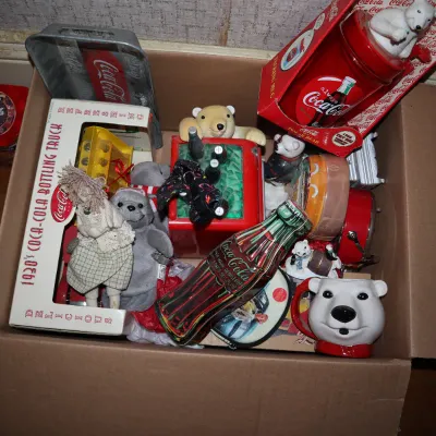 Sale Photo Thumbnail #154: Cardboard box packed with vintage Coca-Cola memorabilia, including a polar bear mug, bottle-shaped tins, a die-cast 1930s bottling truck, and plush toys.