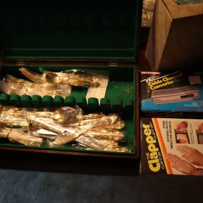 Sale Photo Thumbnail #121: Gold-toned flatware set in a felt-lined wooden storage chest, displayed alongside vintage boxed electronics including a cable channel converter and The Clapper.