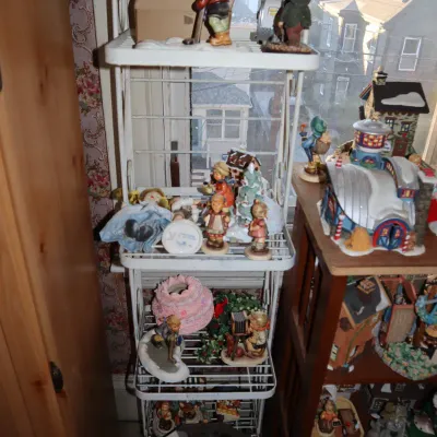 Sale Photo Thumbnail #19: Collection of porcelain Hummel figurines, vintage holiday decor, and miniature village houses displayed on a white wire rack and wooden shelving unit.