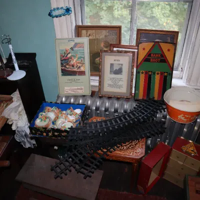 Sale Photo Thumbnail #74: An assortment of vintage items, including plastic toy train tracks, framed artwork and poetry, small teacups in a box, doll stands, and decorative housewares arranged near a radiator.